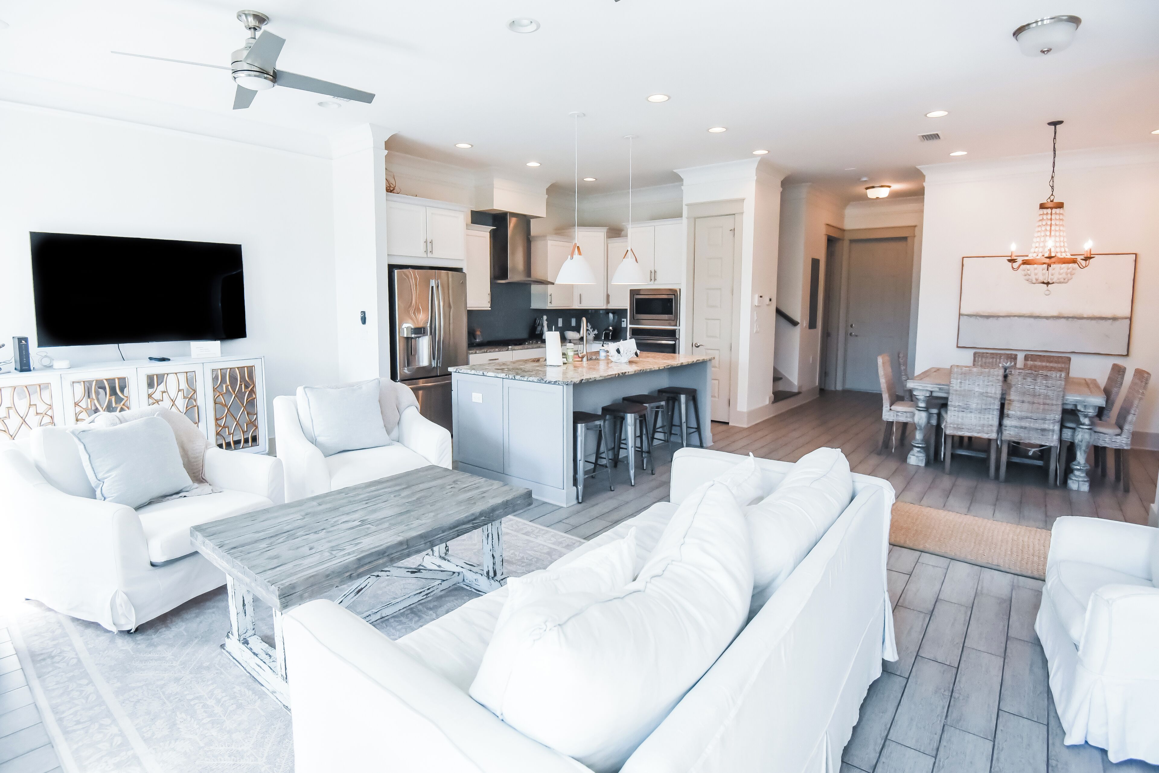 A modern living room with a large TV, white sofas, a wooden coffee table, and a kitchen area with a central island.