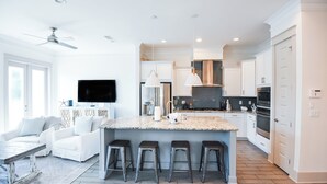 A modern kitchen with white cabinets, a central island with stools, a ceiling fan, and a flat-screen TV mounted on the wall. - Tryin' To Catch A Good Time-30A Townhome with 6-Seater Golf Cart! Sleeps 8 (Santa Rosa Beach)