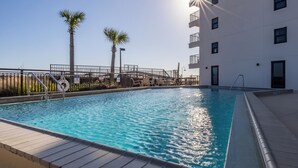 A swimming pool with a clear blue water, surrounded by a white building with balconies, palm trees, and a sun shining from the right. - Introducing Easy Breezey! 3 BR Gulf Front Listing With A Million Dollar View (Pensacola, FL. 32507)