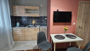 A compact kitchen with wooden cabinets, a black tiled backsplash, a sink, and a small dining table with two chairs. A flat-screen TV is mounted on the wall above the table. - KNEZEVIC RESORTS 1
APARTMANI ZA ODMOR I UZIVANJE (Athina)