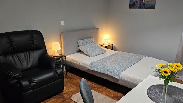 A small, neatly arranged bedroom with a bed, a chair, a nightstand, and a table with a vase of flowers. - KNEZEVIC RESORTS 1
APARTMANI ZA ODMOR I UZIVANJE (Athina)