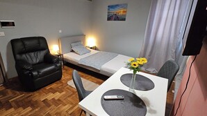 A small, neatly arranged room with a bed, a chair, a table with a vase of flowers, and a TV. - KNEZEVIC RESORTS 1
APARTMANI ZA ODMOR I UZIVANJE (Athina)