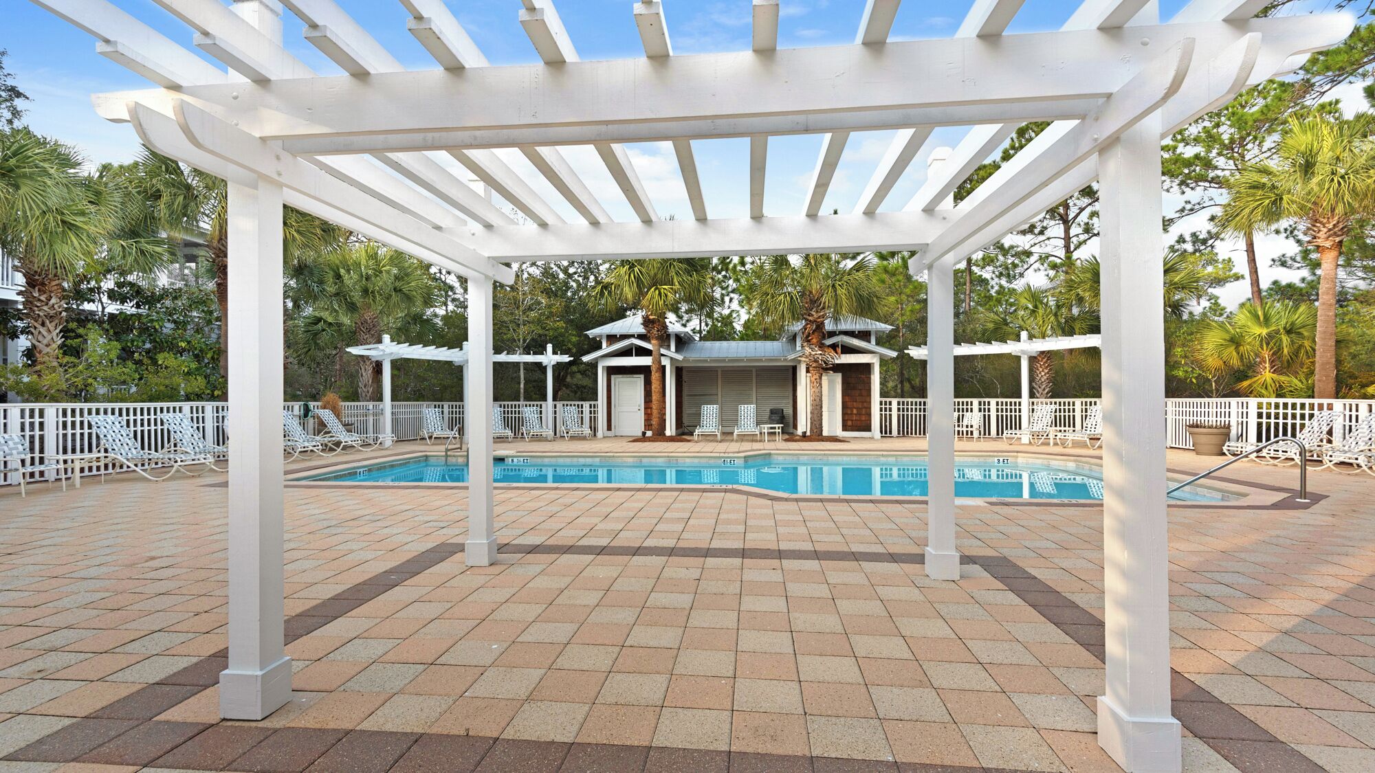 A covered outdoor pool area with white pergolas, lounge chairs, and a small building with a roof.