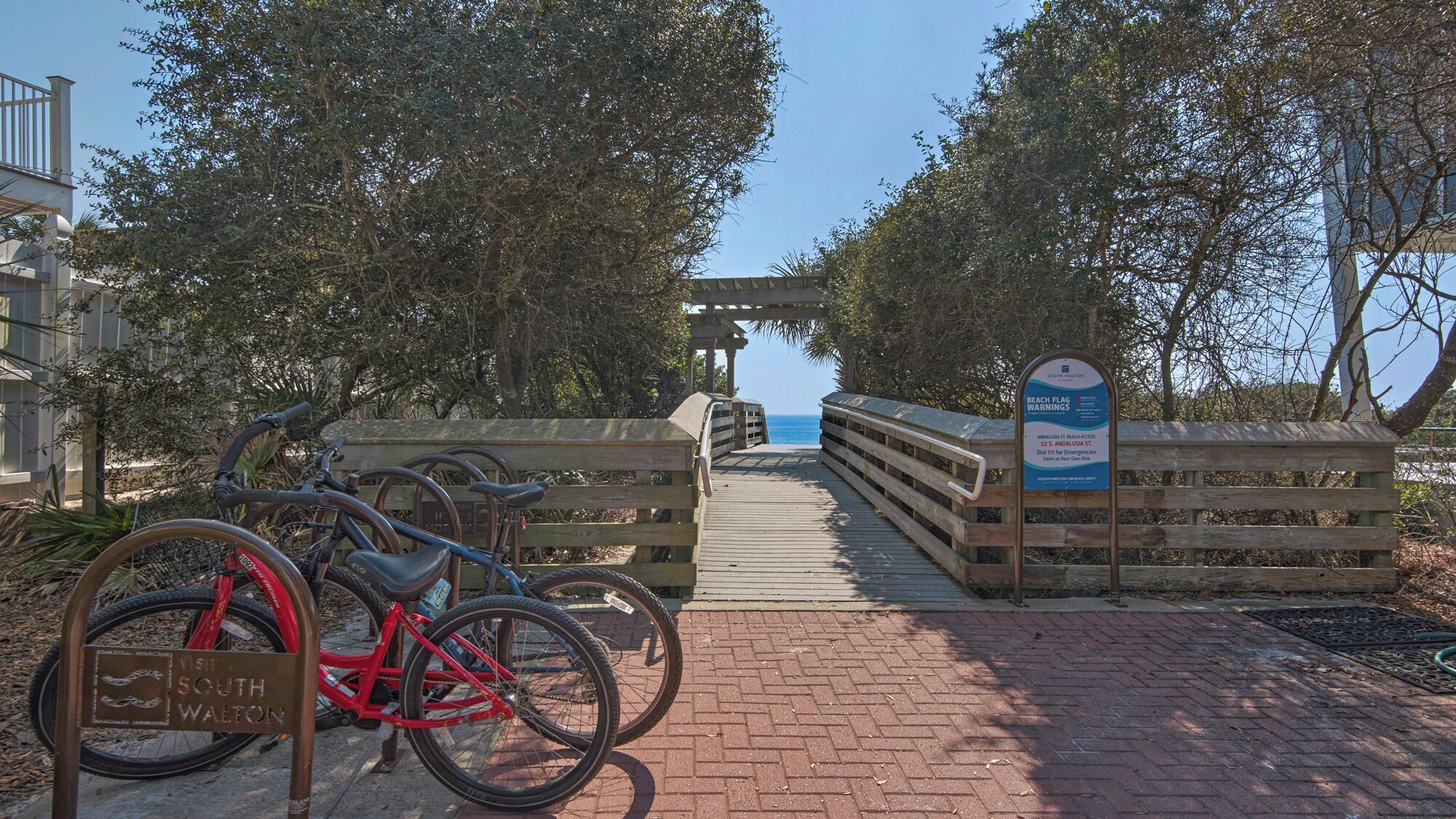 Ein Holzweg führt zu einem Strandabschnitt, wo Warnschilder für den Strand und ein Fahrradständer mit Fahrrädern zu sehen sind.
