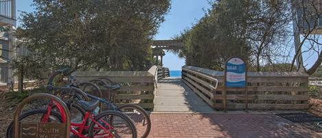 Ein Holzweg führt zu einem Strandabschnitt, wo Warnschilder für den Strand und ein Fahrradständer mit Fahrrädern zu sehen sind.