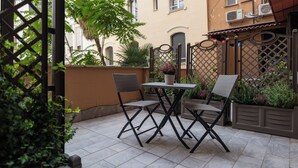 Eine Terrassengruppe im Freien mit Tisch und zwei Stühlen, umgeben von Grünpflanzen und einem Metallzaun.