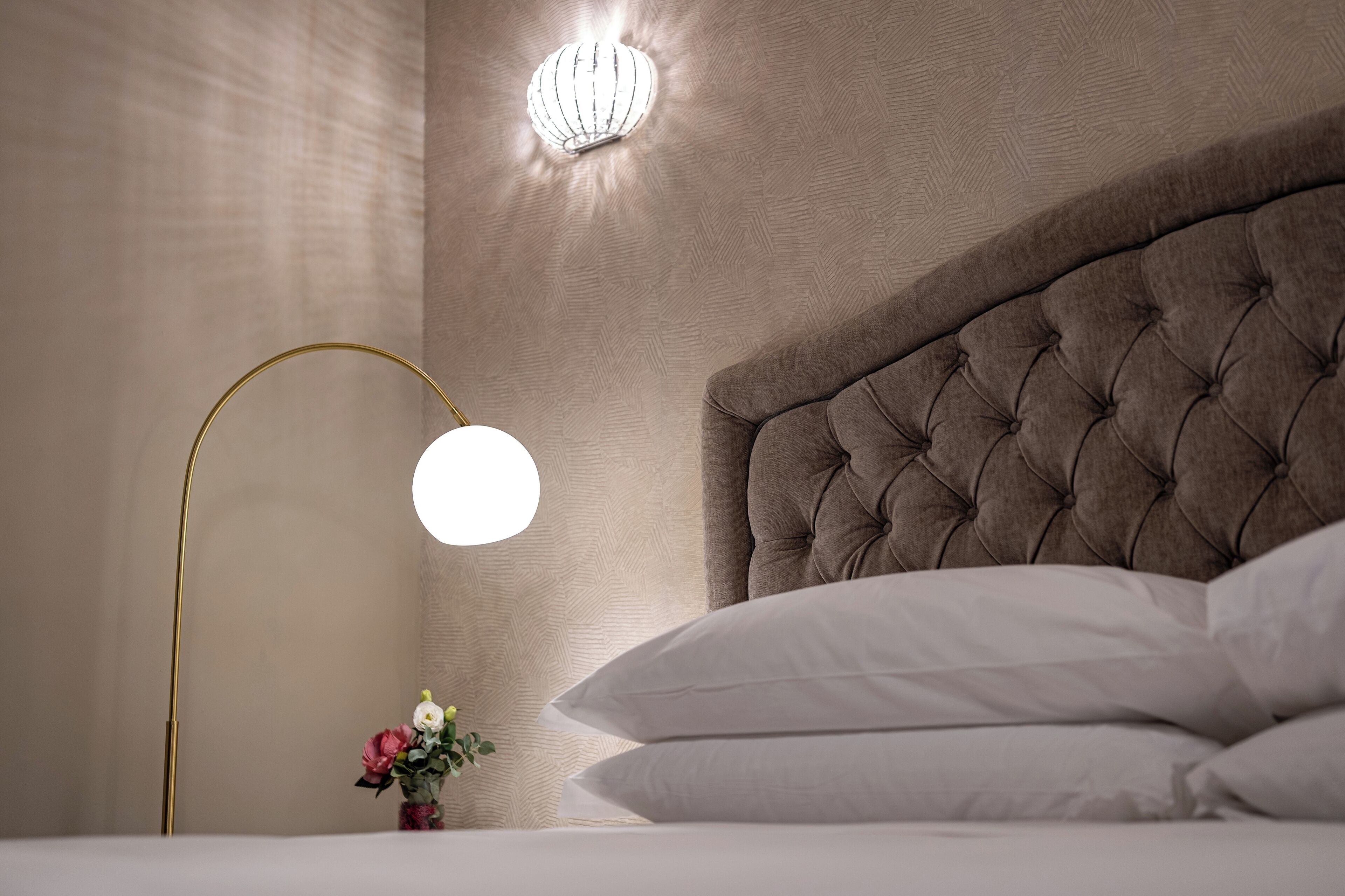 A bedroom with a tufted headboard, a bedside lamp, and a small vase with flowers.