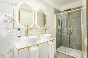 Un baño con dos lavamanos blancos, detalles dorados y una ducha con cerramiento de vidrio.