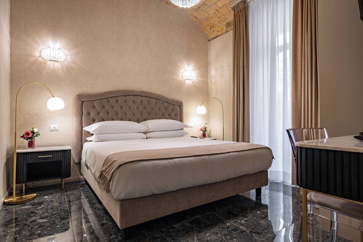 Une chambre avec un grand lit, deux tables de chevet avec des lampes, une petite table avec un vase et une vue sur l’extérieur par une fenêtre avec des rideaux.