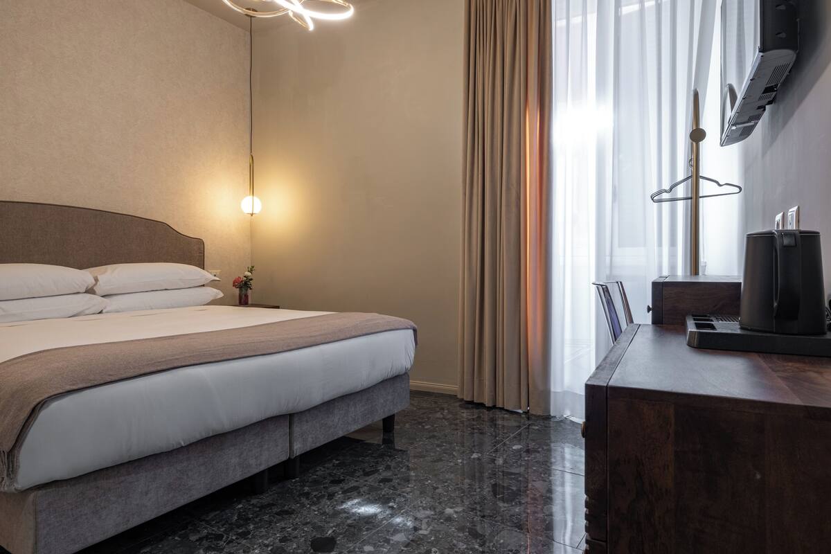 Un lit bien fait, avec une tête de lit, une lampe de chevet, une commode en bois avec une machine à café, et une fenêtre avec des rideaux.