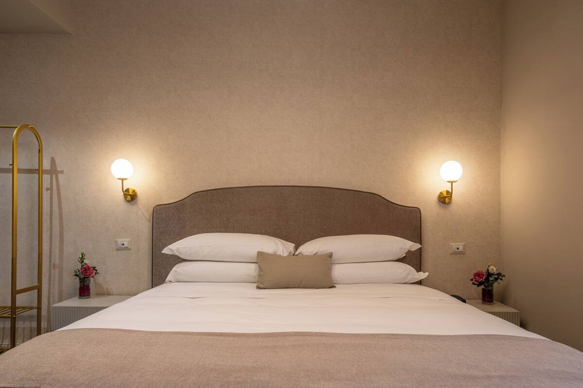 Un lit bien fait, avec deux oreillers, une table de chevet sur laquelle se trouve une lampe, et un petit vase avec des fleurs.