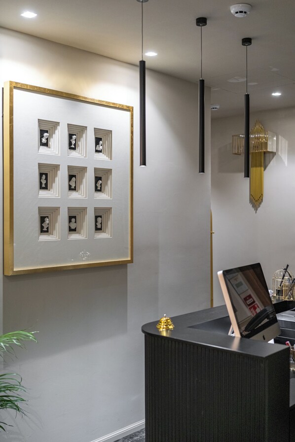 A reception area with a framed artwork of multiple small sculptures, a reception desk with a golden ornament, and pendant lighting. - Roma Suite Hotel (Rome)