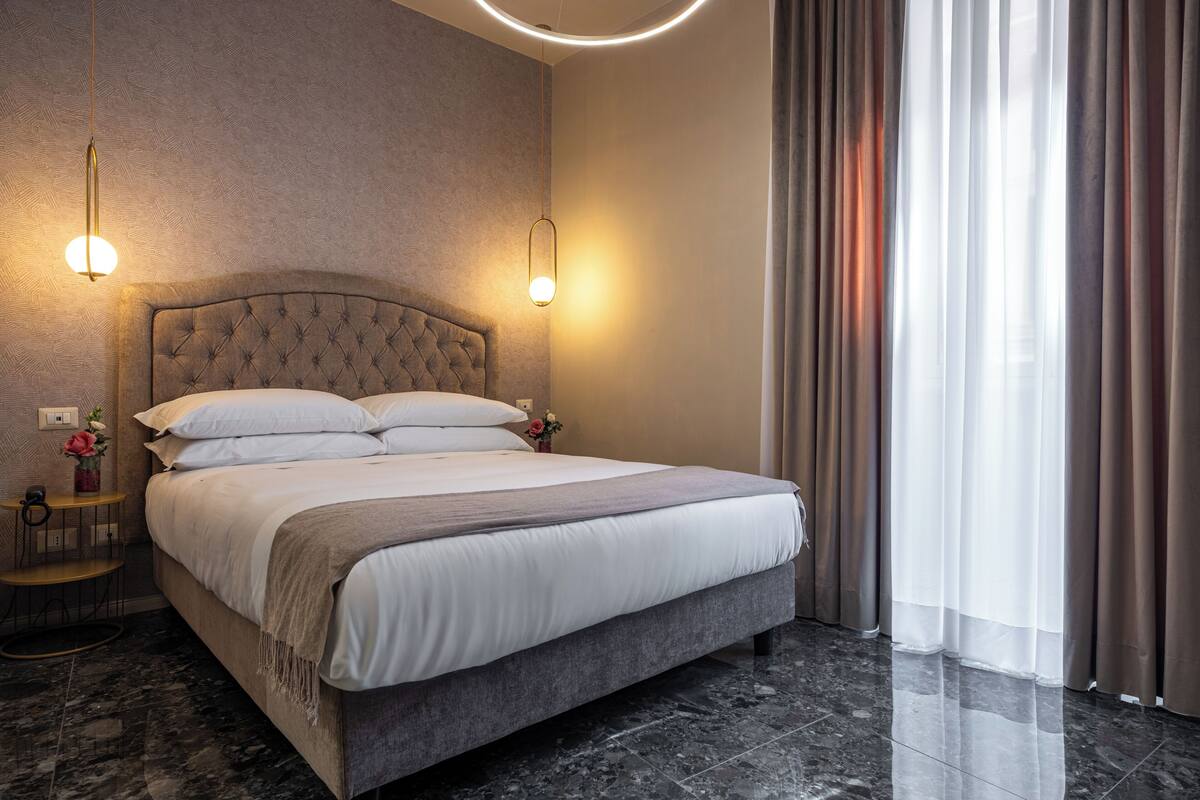 Une chambre d’hôtel moderne avec un grand lit, deux lampes de chevet, un sol en marbre et une vue sur l’extérieur à travers de fines rideaux.