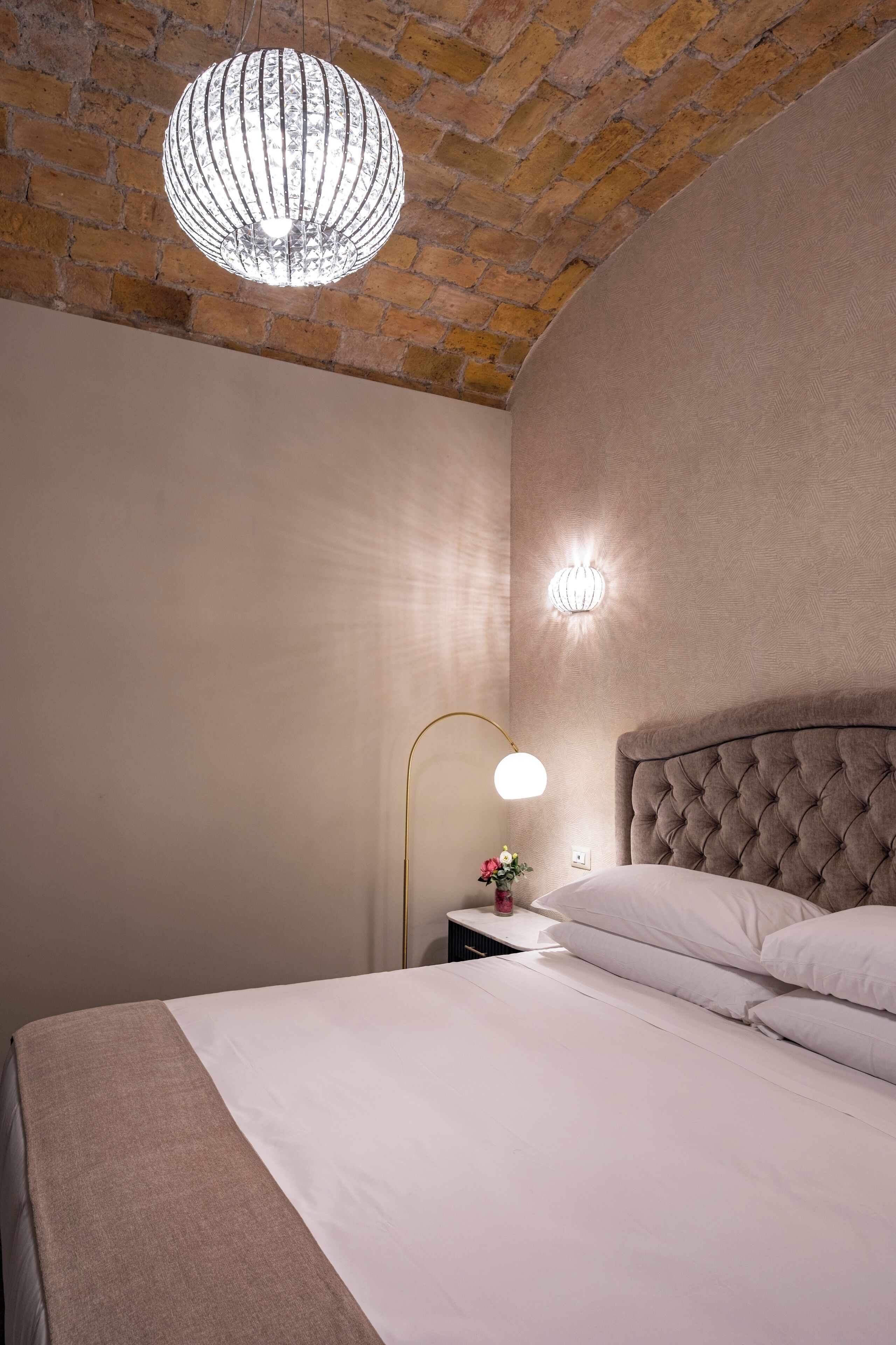 A bedroom with a brick ceiling, a tufted headboard, a bedside lamp, and a wall-mounted light fixture.