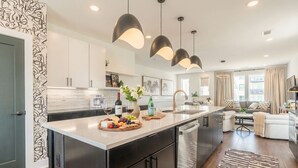 A modern kitchen with a central island, pendant lighting, and a dining area with a sofa and coffee table. - Luxe 4BR With Fire Pit Grill and City Views (Nashville)