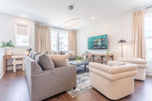 Un salon moderne avec un canapé gris, un pouf beige et une table basse. Un téléviseur est fixé au mur, il y a un bureau avec une chaise et une lampe.