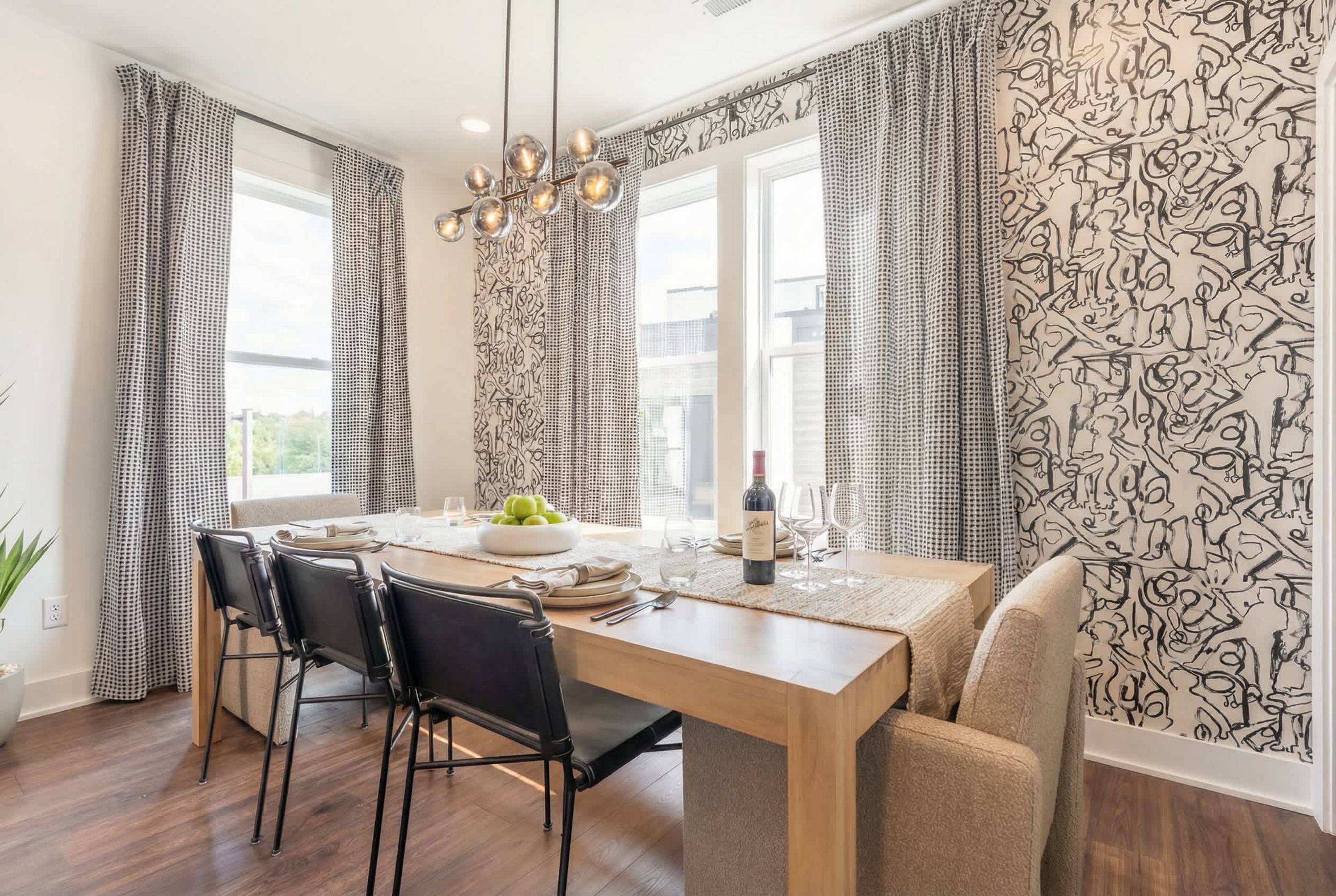 Un comedor con mesa de madera, sillas negras, una araña y cortinas estampadas.