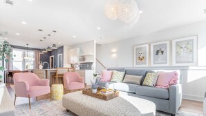 A modern living room with a grey sofa, pink armchairs, and a large ottoman. There is a kitchen in the background with white cabinets and a dining area. - Nashville Retreat With Hot Tub Private Garage (Nashville)