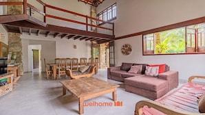 A spacious living area with a high ceiling, wooden furniture, and large windows offering a view of greenery.
