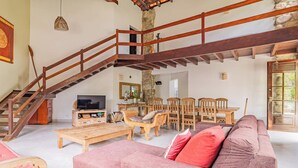 A spacious living area with a wooden staircase, a dining table with chairs, a television, and a large sofa.