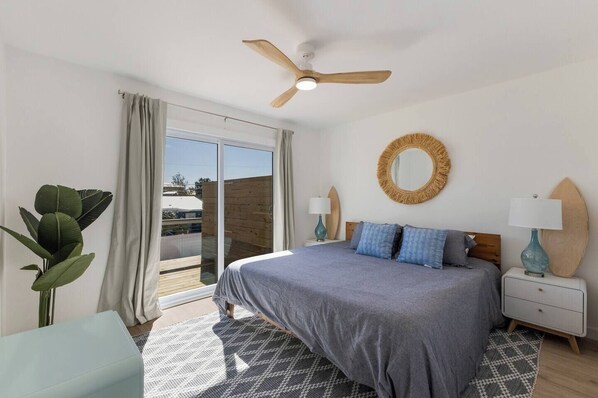 A bedroom with a bed, a ceiling fan, a round mirror, and a surfboard on the nightstand.