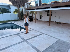 Una piscina con área de deck con baldosas, estructura de deck de madera y una parrilla al fondo.