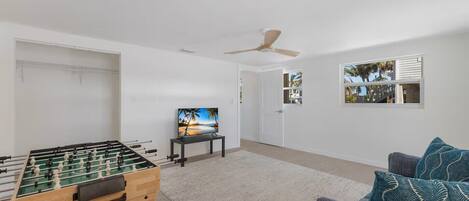 A room with a foosball table, a television on a stand, a ceiling fan, and a window with a view of palm trees.