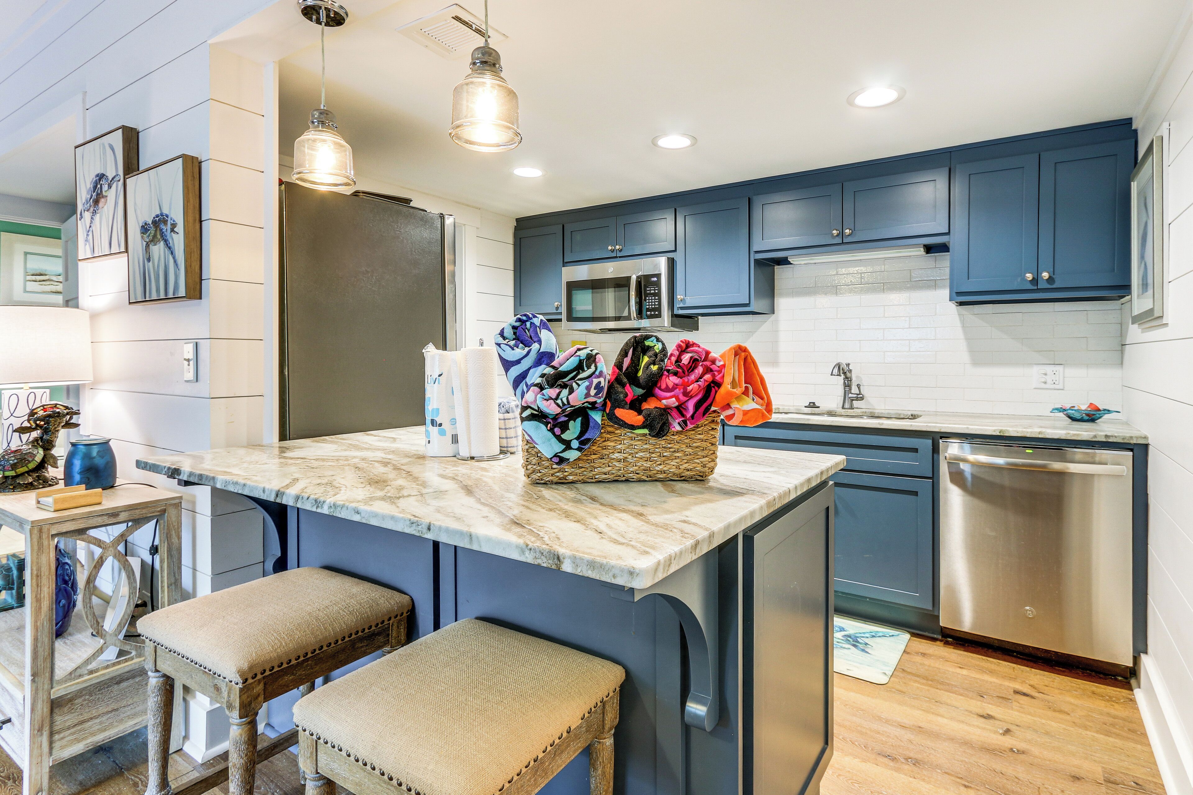 A modern kitchen with blue cabinets, a stainless steel refrigerator, a microwave, and a dishwasher. There are two bar stools and a woven basket with colorful towels.