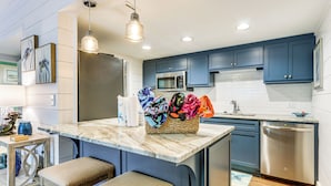 Una cocina moderna con gabinetes azules, un refrigerador de acero inoxidable, un microondas y un lavavajillas. Hay dos taburetes de bar y una cesta tejida con toallas coloridas.
