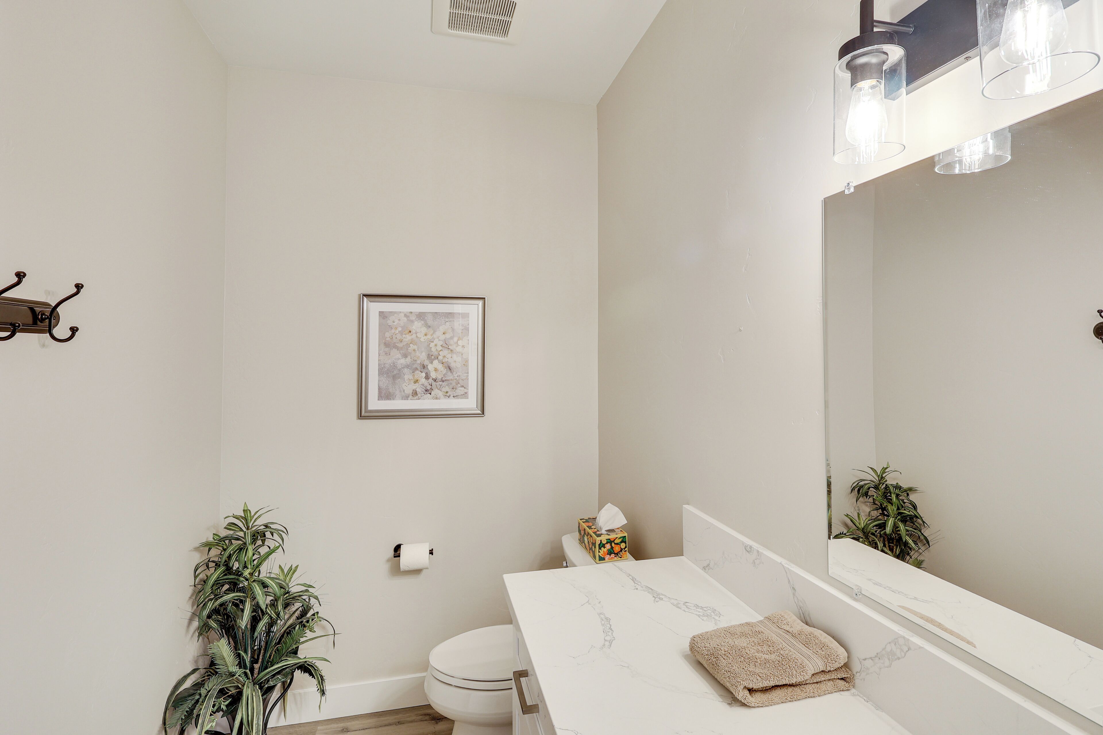 Un bagno con un water bianco, un lavandino con piano in marmo, un asciugamano, un quadro incorniciato appeso al muro e una pianta in vaso.