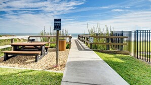 A beachside picnic area with wooden benches and picnic tables, a concrete pathway, and a fenced area with a sign indicating beach access. - Steps to Ocean, Pool Access! Daytona Beach Gem (Daytona Beach Shores)