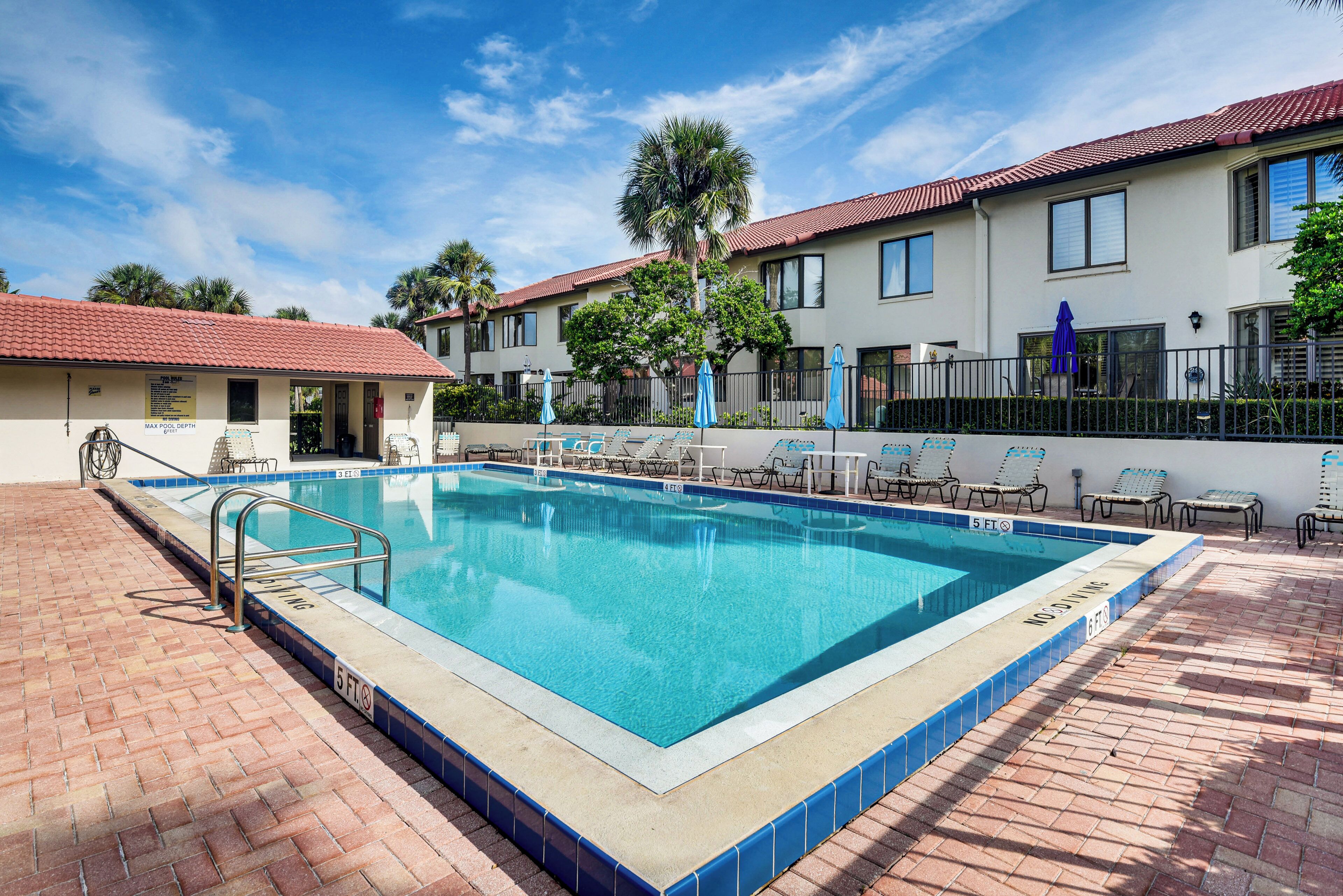A rectangular swimming pool with a tiled edge, surrounded by lounge chairs and umbrellas, with a brick patio and a building with a red-tiled roof in the background.