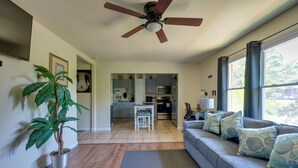 Un salon avec un canapé gris, un ventilateur de plafond, une plante en pot et une vue sur la cuisine par une porte ouverte.