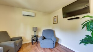 Un salon comprenant un fauteuil gris, un canapé, un téléviseur à écran plat fixé au mur, un climatiseur, une petite table avec une lampe et une grande plante verte.
