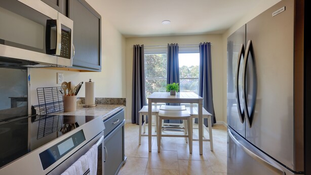 Una cocina moderna con microondas, horno y refrigerador integrados. Hay una mesa de comedor con sillas y una ventana con cortinas.