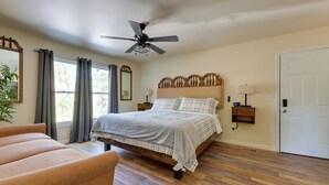 Un dormitorio con cama, ventilador de techo, mesita de noche con lámpara, mesita auxiliar, cabecera de madera, puerta y ventana con cortinas.