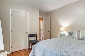 Un dormitorio con una cama, una silla, una mesita de noche con lámpara y una puerta que da a otra habitación.