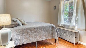 Egy hálószoba, amelyben található egy ágy, egy éjjeliszemélyzet, egy lámpa, egy komód és egy ablak, amelyen függöny van.