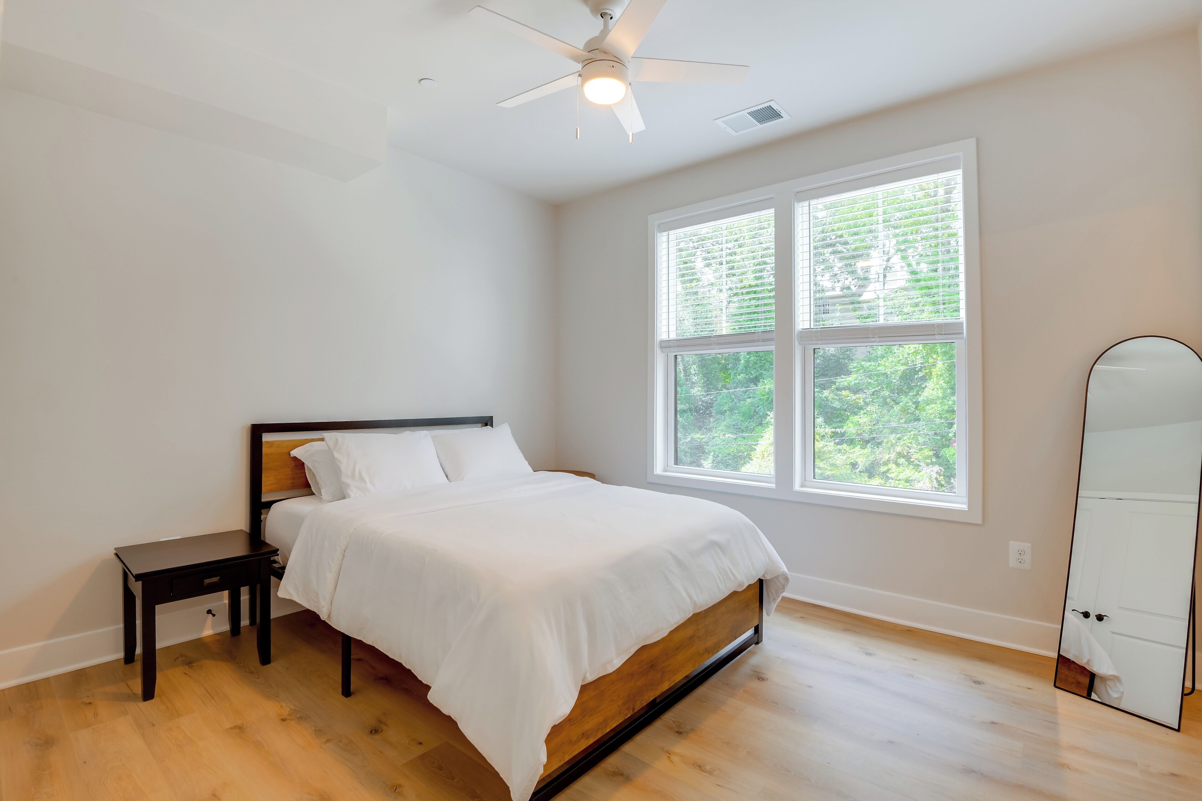 A bedroom with a bed, a nightstand, a ceiling fan, a large window, and a mirror.
