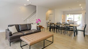 Un salon moderne avec un canapé en cuir foncé, un pouf beige et un coin repas comprenant une table et des chaises en bois.