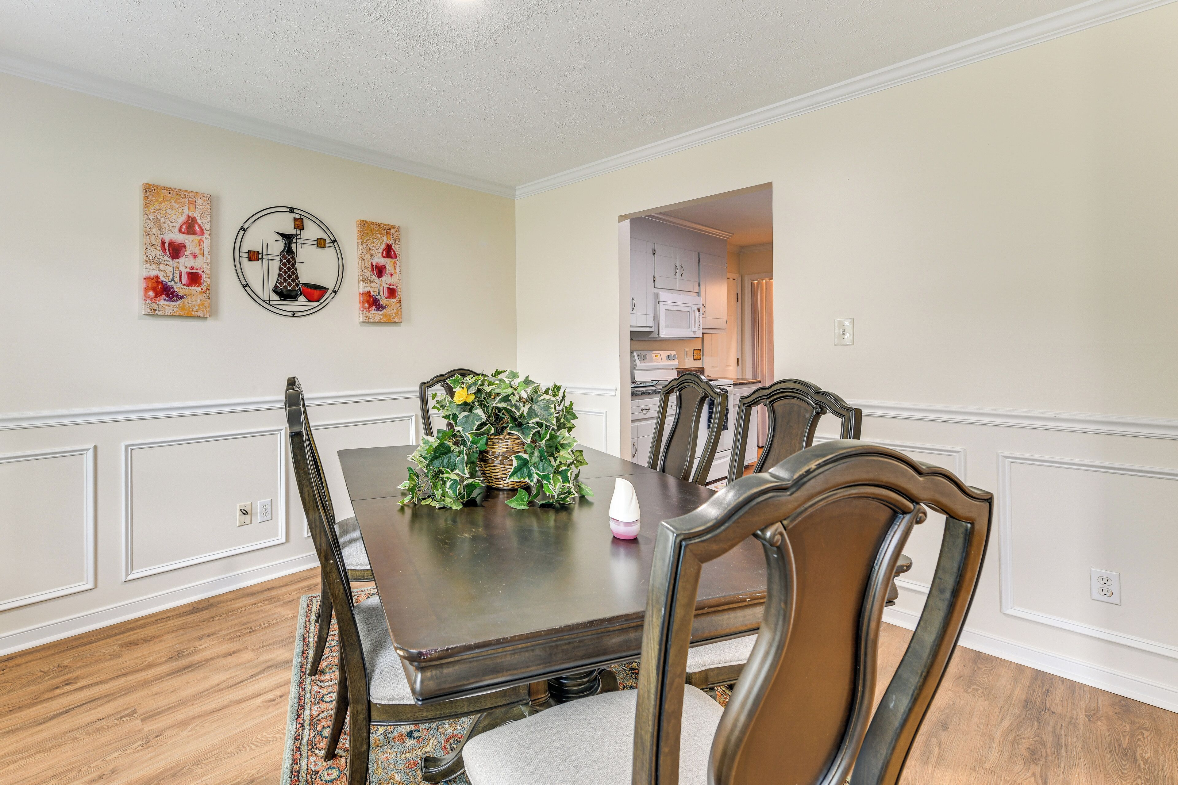 Une salle à manger avec une table en bois, six chaises et un vase rempli de fleurs. Trois tableaux ornent le mur.