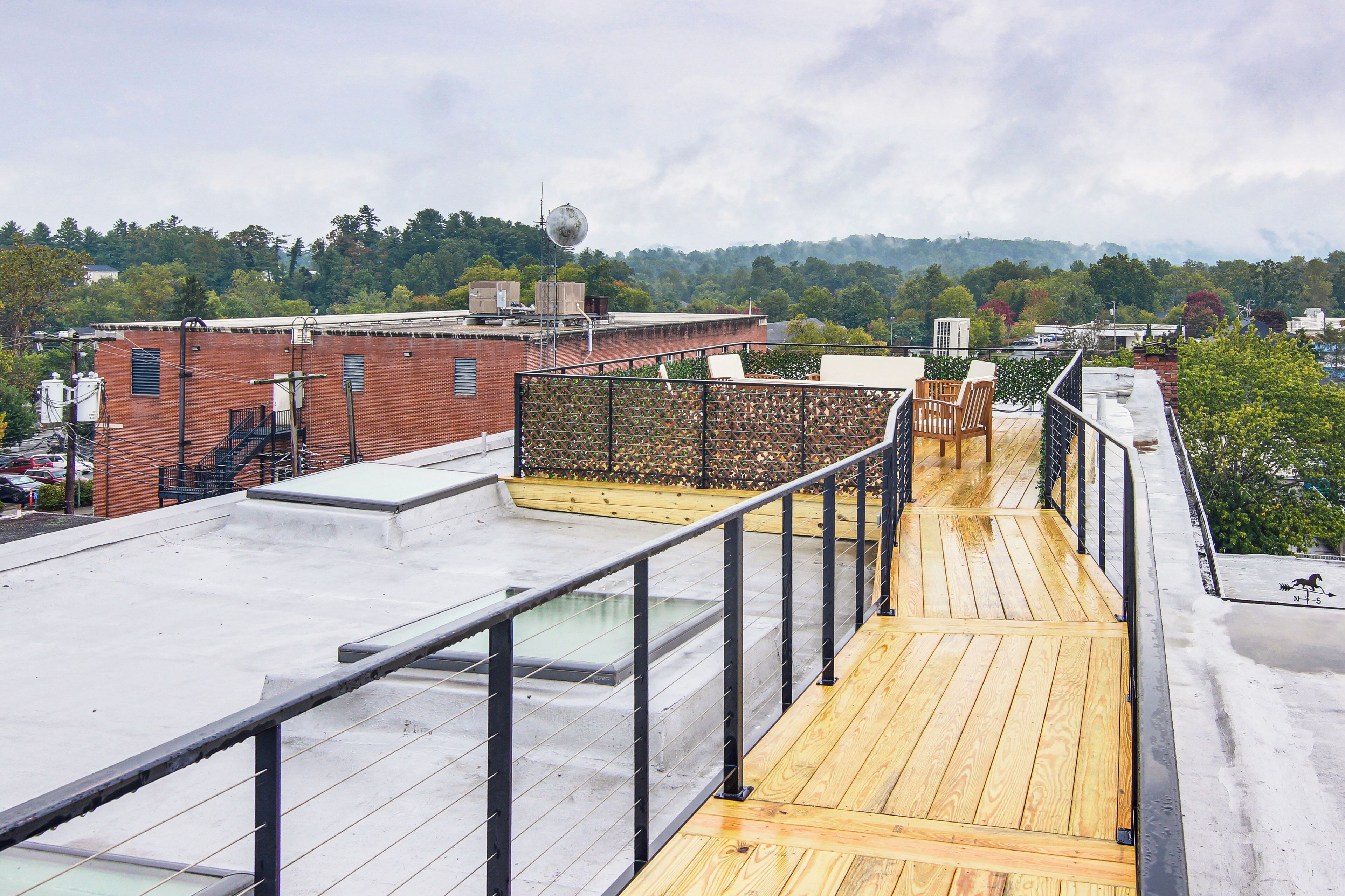 Una terraza en la azotea con piso de madera y barandillas metálicas, con vista a un paisaje urbano con edificios y árboles.