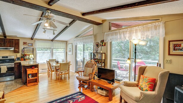 A spacious living area with wooden flooring, a ceiling fan, a fireplace, and a large window offering a view of the outdoors. - Quiet All-season Creekside Escape in Upstate! (Berlin)