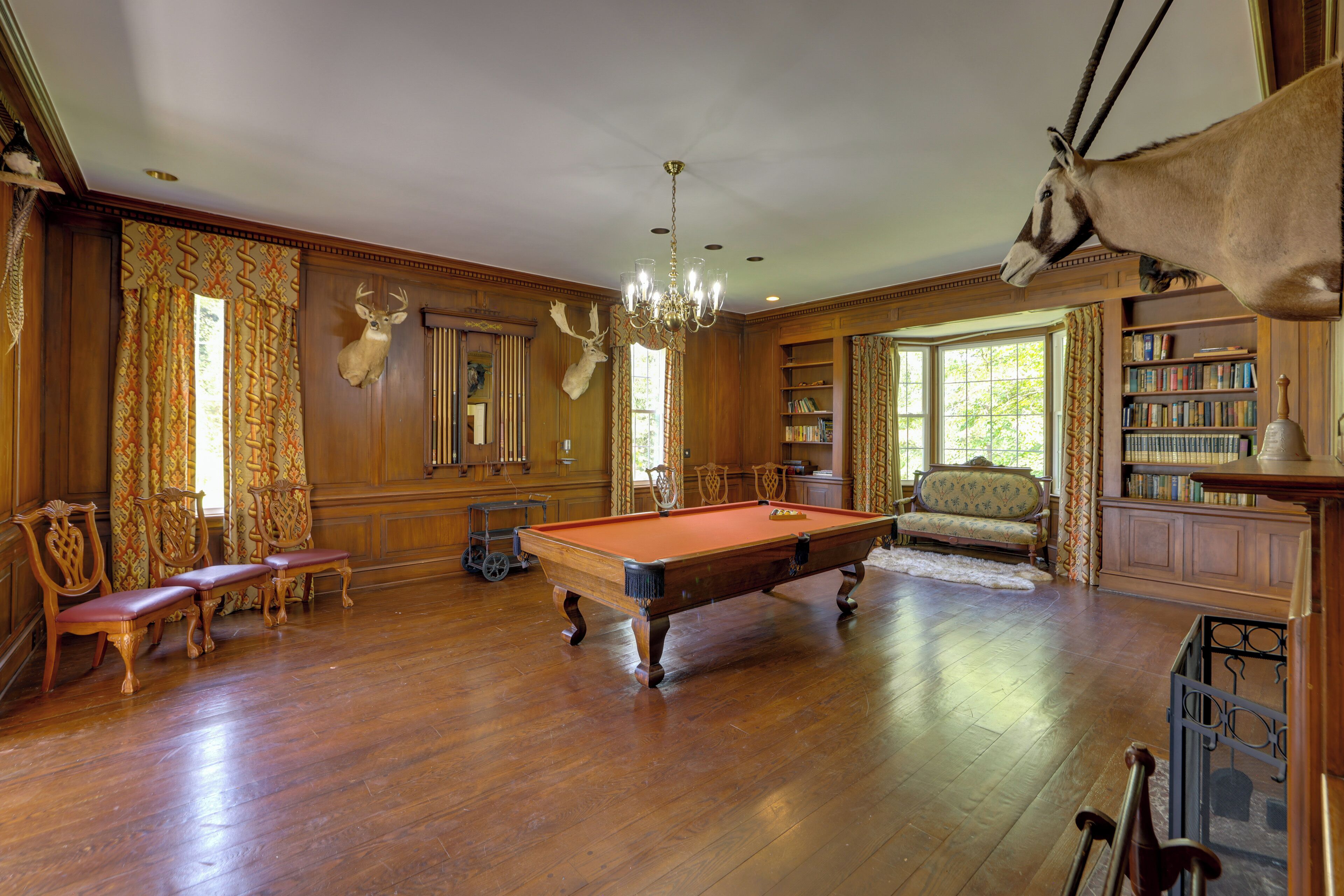 A spacious room with a pool table, wooden furniture, a chandelier, and a deer head mounted on the wall.