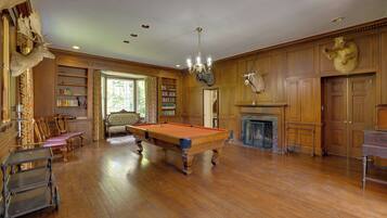 Une pièce spacieuse équipée d’une table de billard, de bibliothèques en bois, d’une cheminée et d’un lustre.