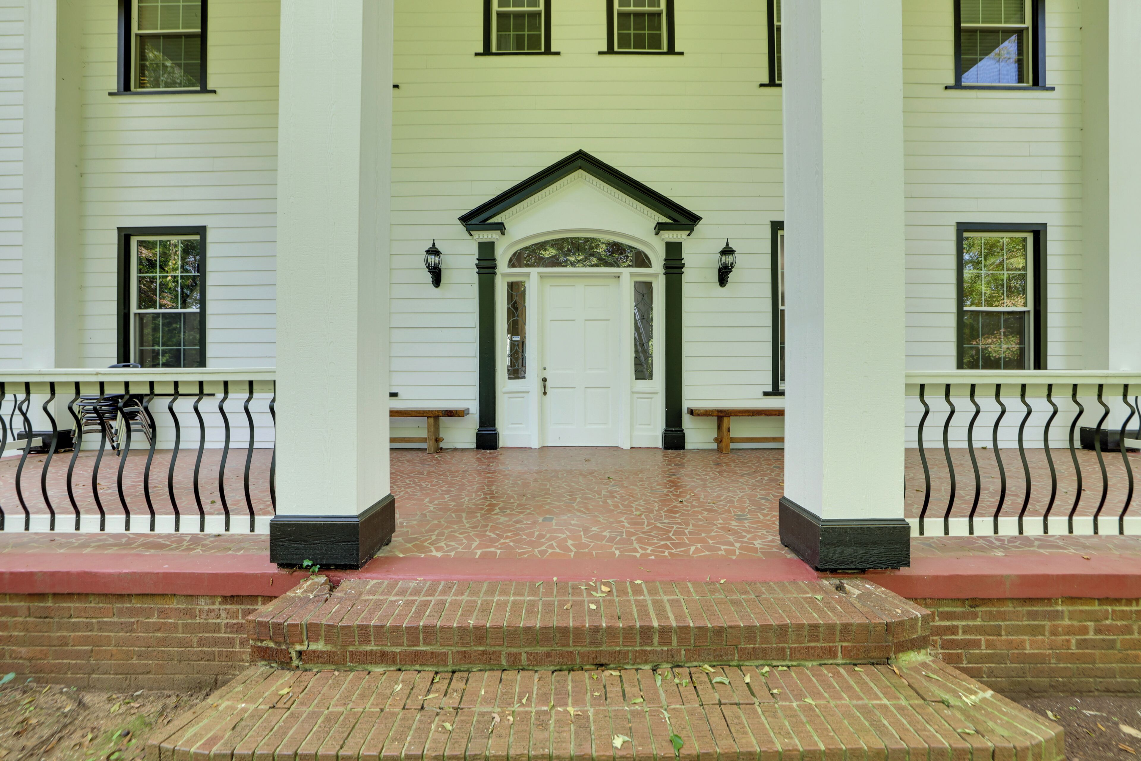 A white building with a central entrance, flanked by two columns and a brick-paved area leading to the door.