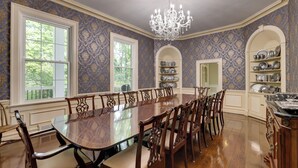 Una sala da pranzo formale con un lungo tavolo in legno e sedie, circondata da carta da parati decorata e scaffali a muro con stoviglie.