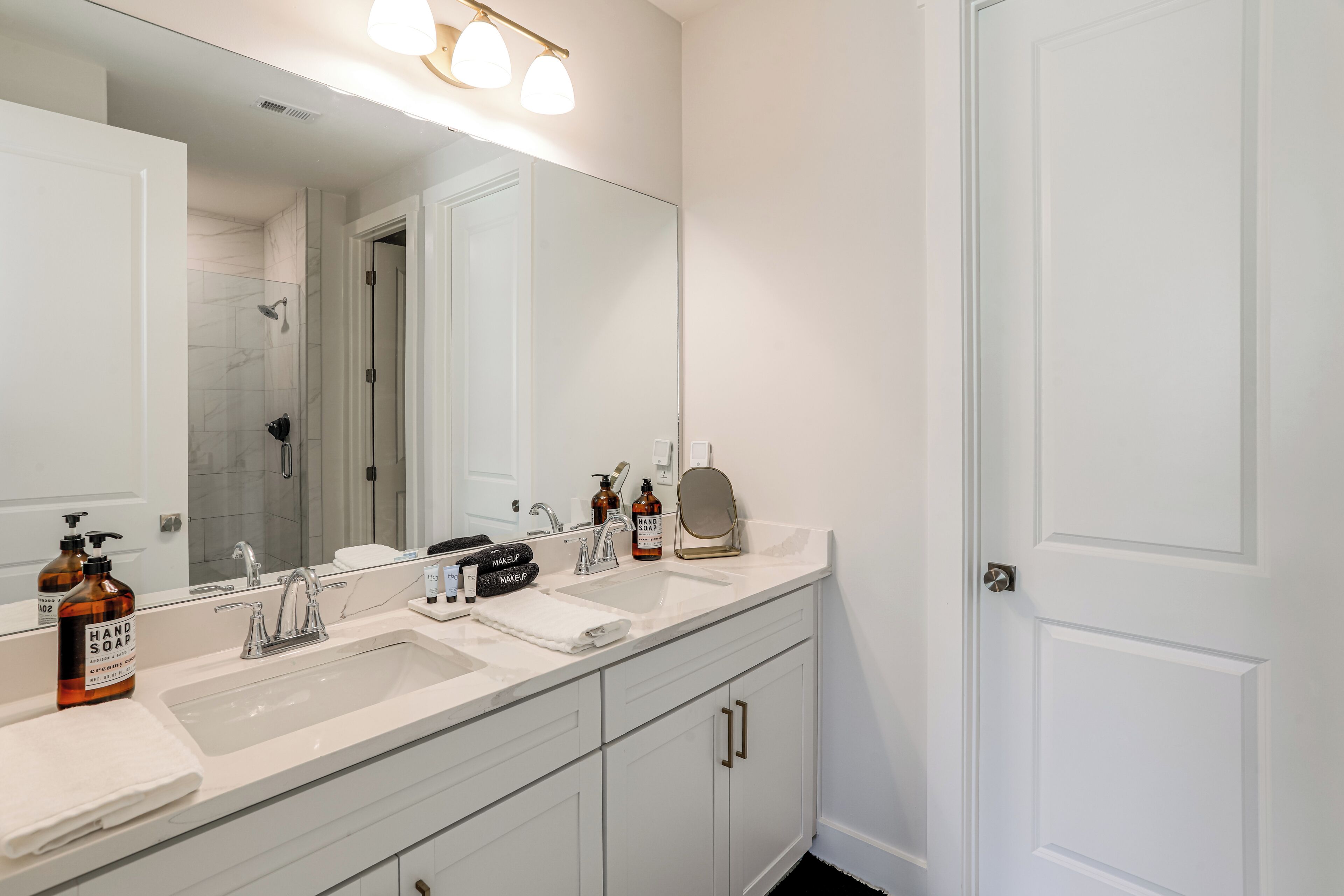 A bathroom with a large mirror, two sinks, a shower, and a vanity with drawers.