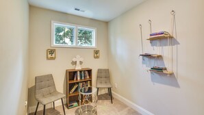 Ein Zimmer mit zwei Stühlen, einer Bücherwand und einem kleinen Tisch. Ein Fenster bietet Blick auf Bäume, und eine Wandregal beherbergt Bücher.