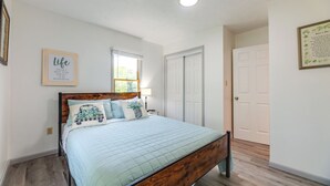 Ein Schlafzimmer mit Bett, Fenster, Tür und einer Wand mit einem gerahmten Zitat.